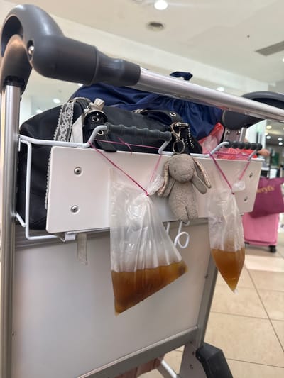 The image depicts the contents of a luggage cart in an indoor setting, likely within an airport in George Town, Malaysia, based on common local practices of carrying drinks in plastic bags. The cart holds a blue backpack, a black purse with white lace trim, a stuffed rabbit, and two plastic bags containing a brownish liquid, presumably drinks, secured with rubber bands to the cart's structure. The cart itself is white and silver, with a gray handle. The background shows glimpses of a tiled floor and a pink suitcase, suggesting a baggage claim area or a similar transit space.