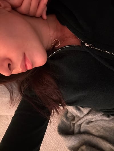 A person is lying down, facing upwards. The face is visible from the nose to the forehead, with lips slightly parted. A silver necklace and earring are visible on the neck. The person is wearing a black zip-up jacket. Auburn hair is spread around the head. Part of a hand is visible near the neck. A gray blanket or cushion is visible in the background. The overall lighting appears dim.