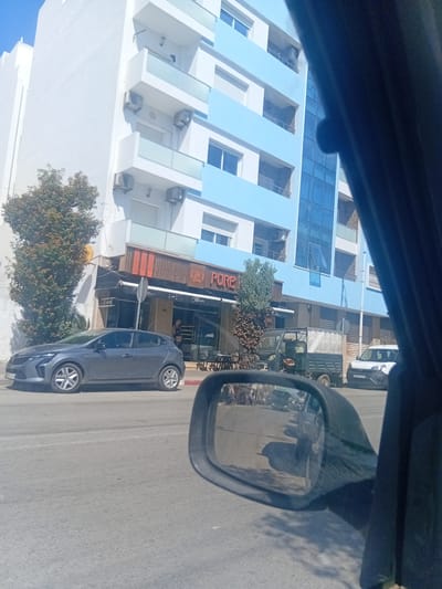 The image, taken from inside a vehicle, shows a street scene in Martil, Morocco. A light blue and white building with multiple windows stands along the street. A cafe called "Pure" is also visible, with signage indicating "Cafeteria, Patisserie, Boulangerie". A gray car is parked on the side of the road. A vehicle similar to a pickup truck and a white van are visible further down the street. A side mirror is in the foreground, reflecting the surrounding scene. Trees are present on the roadside.