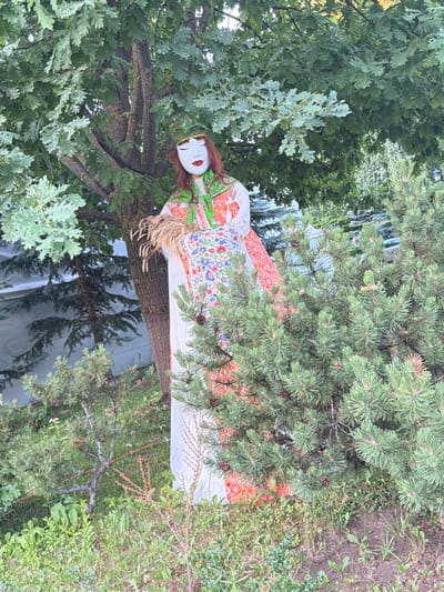 A full shot captures a life-sized effigy of a woman in traditional attire, positioned outdoors amidst lush greenery. The effigy, draped in a white gown adorned with vibrant floral embroidery and a decorative green collar, stands tall. A golden, jeweled headdress rests upon its head, accentuating a white mask-like face with striking red lips and closed eyes. Its right arm clutches a bundle of dry wheat stalks. The effigy is partially obscured by the branches of a large oak tree overhead and dense pine shrubs to its right. The background reveals a building with a textured, light-colored facade and hints of other foliage. The ground is covered with a mix of green grass and dry soil, with small plants and twigs scattered throughout. The scene appears to be a public space, possibly a park or garden, in the city of Suzdal, Russia, given the context.