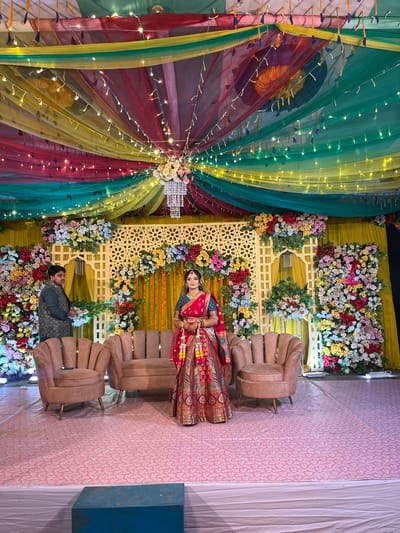 The image displays an elaborately decorated indoor event venue, located in Savar, Bangladesh. A stage is set with a backdrop of yellow curtains overlaid with ornate white lattice panels, heavily adorned with multi-colored artificial floral arrangements in hues of red, pink, yellow, white, blue, and green. The ceiling is draped with translucent fabrics in red, yellow, green, and teal, illuminated by numerous strands of fairy lights and a central chandelier decorated with white and pink flowers.
Two individuals are present on the stage. A female subject stands centrally, facing the viewer, dressed in a red lehenga with gold embroidery, a teal blouse, and a red dupatta. She wears extensive traditional jewelry, including a maang tikka, necklace, earrings, and bangles, and holds colorful floral garlands in her clasped hands. To her left, slightly behind her, a male subject stands facing right, wearing a dark textured jacket over a dark shirt, with his hands clasped in front.
Three pieces of light brown or blush pink upholstered furniture, comprising two armchairs and one sofa with ribbed backs and brass-colored legs, are arranged on the stage. The floor is covered with a light pink carpet featuring a subtle white floral pattern. A rectangular blue block rests on the carpet in the foreground.