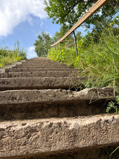 The image depicts an upward perspective of a weathered, concrete outdoor staircase. The steps are rough and worn, with visible texture and debris. Lush green grass and vegetation grow along the sides and between the steps, suggesting an overgrown or naturalized environment. A wooden handrail, supported by dark metal posts, runs along the right side of the staircase, angling upwards and away from the viewer. In the background, trees and a bright blue sky with scattered white clouds are visible. The overall impression is one of an aging, naturalized outdoor space, possibly a park or pathway. The location is identified as Cekule, Latvia. There are no people or animals present in the image, and no specific actions or interactions are captured.
