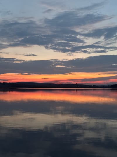 A wide-angle, vertical photograph captures a serene sunset over a calm body of water, likely a lake or a wide river, in Berezhany, Ukraine. The sky is a gradient of colors, transitioning from a pale blue at the top to a vibrant orange and red band near the horizon. Wispy, dark grey clouds are scattered across the upper portion of the sky.

A silhouetted, low-lying treeline or hills form a dark, continuous line along the horizon, separating the water from the sky. The water itself is remarkably still, acting as a mirror that reflects the fiery hues of the sunset and the muted tones of the clouds. These reflections are slightly distorted by gentle ripples on the water's surface, creating an abstract pattern of color and light.

The overall impression is one of tranquility and natural beauty, with no human figures or man-made structures prominently visible. The scene is entirely dominated by the interplay of light, color, and reflection in the natural environment.