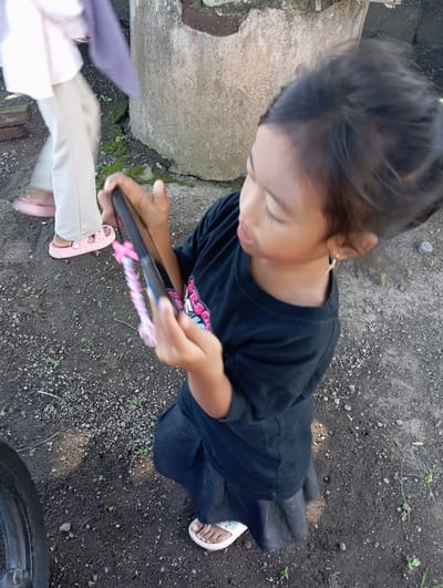 A young child stands on a dirt ground with sparse vegetation, possibly in a semi-urban area, holding a mobile device in their hands. The location is likely in Waung, Indonesia. The child is wearing a dark-colored shirt and appears to be focused on the screen. Another person's legs and feet, wearing light-colored pants and pink sandals, are visible in the lower-left corner, suggesting they are standing nearby. A concrete structure is visible on the left side of the frame. A portion of a tire is visible in the bottom right of the frame.