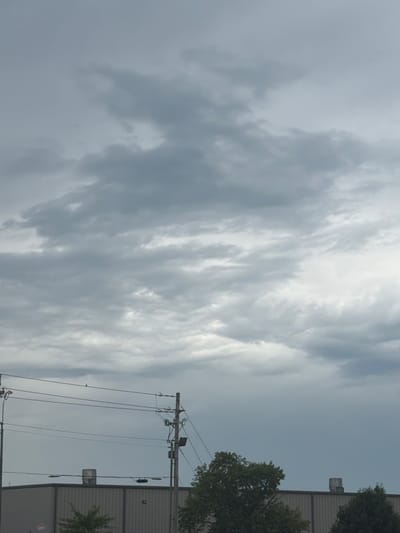 The image shows a cloudy sky over a section of Murfreesboro, United States of America. In the foreground, a grey, industrial-style building with a corrugated metal facade is visible. On top of the building, there are two cylindrical vents. Utility poles with multiple wires and streetlights run horizontally across the frame above the building. A few trees with green foliage are positioned in front of and to the side of the building. The sky is overcast with varying shades of grey clouds.