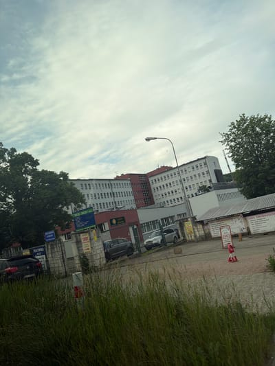 The image shows a street view of buildings in Limanowa, Poland. A large, multi-story building complex is visible, with a white facade and numerous windows. In front of it, there are smaller buildings and a fence. Several signs are posted, including one that reads "SKLEP 1001 DROBIAZGOW", indicating a store selling small items. There are cars parked in the area and tall grass along the roadside. The sky is cloudy.