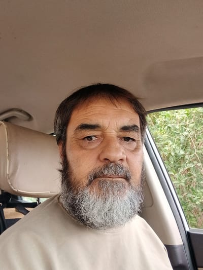 A medium shot photograph shows a man with a grey and brown beard and mustache, and dark hair, looking directly at the camera. He is wearing a tan t-shirt and is seated in the driver's seat of a vehicle. The interior of the car is visible, with a tan headrest to the left of the man. Outside the open car window to the right, green foliage is visible. The lighting suggests it is daytime. The location is identified as Hun, Libya.