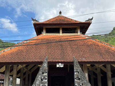 This is a low-angle, eye-level, outdoor, medium shot of a building in Sidemen, Indonesia. The building has a red tile roof and is supported by large pillars. Intricate carvings are visible on the entrance. The sky is blue with some clouds. Power lines are strung across the top of the image. The building appears to be a temple or traditional Indonesian structure. There are no people visible in the image.