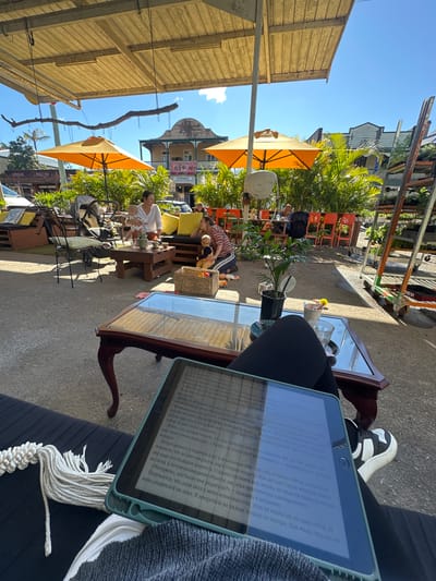 In Bellingen, Australia, a scene unfolds under a covered outdoor seating area, likely a cafe or restaurant. Sunlight filters through gaps in the wooden roof and casts shadows on the concrete ground. Two women are present: one, holding a baby, sits on a yellow cushioned bench with a small table in front of her. The other woman, with dark hair, is seated opposite, looking towards the first woman. A stroller is nearby. In the foreground, a person is seated, with their legs crossed, holding a tablet computer. The tablet displays text from a book. A decorative white macrame piece hangs to the left of the person. A dark wooden coffee table with a glass top is positioned between the foreground person and the seated women. Behind the seated women, an array of orange chairs and lush green plants are visible. In the distance, a multi-story building with a distinctive architectural style stands, featuring balconies and awnings. The overall atmosphere suggests a relaxed, daytime gathering.