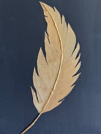 This is a close-up, overhead shot of a textured, gold-colored feather artwork mounted on a dark blue canvas. The feather is centrally placed and oriented diagonally from the bottom left to the top right of the frame. The artwork exhibits a distinct relief, with the feather raised from the canvas surface, creating shadows and highlights that emphasize its textured lines and form. The canvas itself has a subtle woven texture. The image does not depict any specific location in Bevern, Germany, or any people, actions, or interactions.