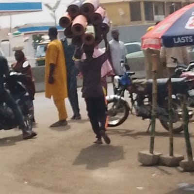The image depicts a street scene in Oko Erin, Nigeria. In the foreground, a person wearing a dark shirt and dark trousers walks towards the right, balancing a tall stack of approximately 15-20 rolled mats or textiles of varying colors on their head, with their arms raised to stabilize the load. To the left of this central figure, a man in a bright yellow traditional tunic and trousers stands. Further left, a woman in a dark red garment is visible with an object on her head. In the mid-ground to the right, a man in a light-colored shirt stands near a parked motorcycle. Another motorcycle is partially visible on the far left. On the right side, a red and blue striped market umbrella is erected, with three concrete-based wooden poles standing in the lower right foreground. The ground is unpaved dirt. In the background, buildings are visible, including one with windows and a structure resembling a gas station canopy on the far left. Several other indistinct figures and vehicles are present in the background. Shadows are cast on the ground, indicating daylight conditions.