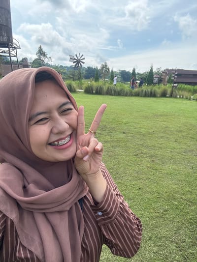 A woman in a brown hijab and striped shirt smiles with a peace sign against a sunny backdrop. Braces are visible on her teeth. Behind her stretches a green lawn, punctuated by trees and distant buildings. A windmill structure is visible to the left. The sky is blue with scattered clouds. The photo appears to be a selfie.