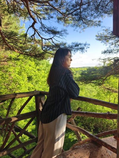 A woman stands on a wooden balcony, looking out at a dense forest under a clear blue sky. The balcony is constructed from rough, natural wood, with branches forming a rustic railing. The woman is wearing a dark pinstripe shirt and light-colored pants. She is seen from the side, with her arms resting on the railing. The background is filled with vibrant green foliage of trees, with hints of distant rolling hills. The scene appears to be set in a natural, outdoor environment, possibly a park or a recreational area in or around the city of Onochino, Russia.