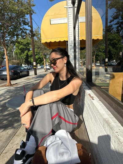 A young female with dark hair and sunglasses is seated outdoors on a concrete ledge next to a building with a large window and a yellow retractable awning. She is wearing a black sleeveless crop top, gray wide-leg pants with red side stripes, white socks, and black and white sneakers. Her left arm is resting on her bent knee, revealing a beaded bracelet and rings on her fingers. A silver necklace and small earrings are visible. To her left, a round grey outdoor table holds a cup with a dark beverage and a pink straw. A brown bag or backpack with a white garment draped over it rests on the ground near her feet. The background reveals a sunlit street scene in Glendale, United States, with a paved sidewalk, trees, utility poles, and parked vehicles, including a dark-colored Tesla Model 3. The sky is clear blue.