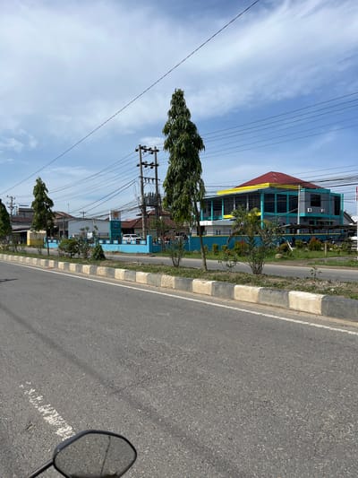 The image depicts a street scene in Tanjungkarang, Indonesia, under a partly cloudy sky. A paved road occupies the foreground, with a white and grey concrete barrier running along its edge. Beyond the barrier, a row of trees and shrubs separates the road from a blue wall.

Behind the wall, a complex of buildings is visible. One prominent building has a red, pyramid-shaped roof with yellow trim and turquoise-colored window frames and vertical accents. This building appears to be a commercial or public establishment, with air conditioning units mounted on its exterior. Another building to the left is mostly white with a blue lower section and a yellow accent. A tall, slender pine tree stands in front of this building. Further down the street, a utility pole with numerous wires is situated.

In front of the buildings, a white car is parked, and several motorcycles are visible. A sign with the acronym "PLN" in blue and white lettering is displayed on a post, suggesting a connection to the Indonesian state electricity company. The overall environment appears to be a developed urban or suburban area, with a mix of architectural styles and infrastructure. The image is taken from the perspective of someone on a vehicle, as indicated by the reflection in a rearview mirror at the bottom left of the frame.
