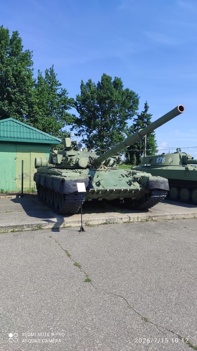 The image shows a static display of two military tanks outdoors under a clear blue sky. The foreground tank is a large, green armored vehicle with its main gun pointed upwards and to the right. It has tracks and a detailed turret with various hatches and equipment. A small white sign is positioned in front of the tank on a black stand. To the right, and slightly behind the first tank, is the rear portion of a second green tank with the number "676" visible on its turret. Behind the tanks, there is a low, green building with a peaked roof and a chain-link fence in front of it. Beyond the building and tanks, numerous tall, green trees are visible, suggesting a park or outdoor museum setting. The ground is paved with asphalt, showing some cracks and sparse vegetation. The overall scene is calm and still, with no people present.