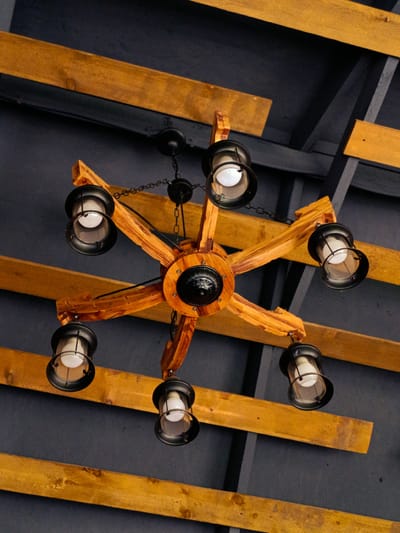 An overhead shot shows a rustic, wooden chandelier hanging from a dark ceiling with exposed wooden beams. The chandelier has a central wooden hub with six arms extending outwards. Each arm supports a black metal lantern with a clear glass shade, containing a visible light bulb. The wooden beams on the ceiling are rough-hewn and have a warm, natural finish. The scene appears to be indoors, likely in a restaurant or a space with a rustic aesthetic. The exact location in Kok Jar, Kyrgyzstan cannot be determined from the visual information provided.