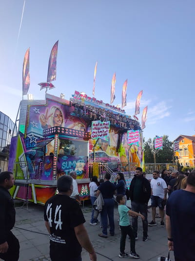 The image captures a vibrant scene at what appears to be a fair or carnival in the city of Bozhurishte, Bulgaria. The focal point is a large, brightly colored amusement ride adorned with flags and images of a woman giving a peace sign, along with the words "Miami Show" and "Summer Times." The ride is in operation, with people seated and enjoying the experience.
A crowd of people, including men, women, and a young boy, are gathered around the ride. The boy is gesturing with his arms. The majority of people are dressed casually. The setting suggests an outdoor event with buildings and trees visible in the background. A contrail streaks across the clear sky.