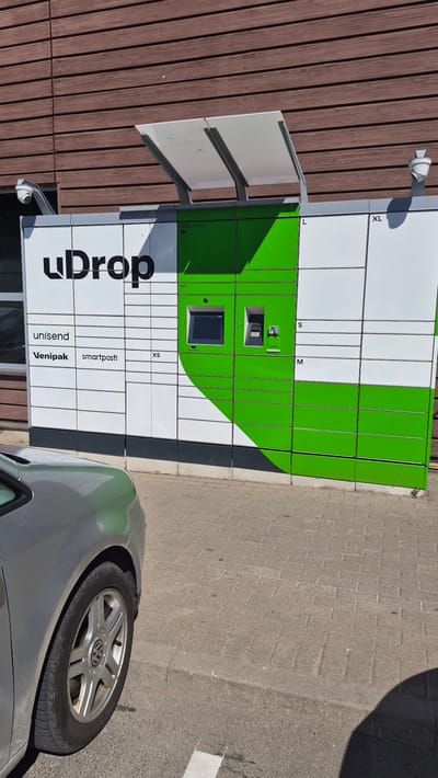 The image shows a UDrop parcel locker station on a sunny day, located in Līvāni, Latvia. The station is a white and green modular unit mounted on a wall with brown wooden siding. The UDrop logo is prominently displayed in black on the white section of the locker. The locker station has various compartments labeled with sizes such as XS, S, M, L, and XL. In the center of the white section, the words "unisend", "Venipak", and "smartposti" are visible. There is a touch screen interface and a card reader on the green section of the locker. Two security cameras are mounted above the locker station. In the foreground, a portion of a silver car is visible on the left side of the frame, with its front tire and wheel clearly in view. The ground in front of the locker station is paved with grey bricks, and a concrete curb separates the paved area from an asphalt surface.