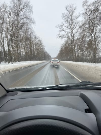 The image depicts a winter road scene in Podmosh'ye, Russia, viewed from the interior of a moving vehicle. The foreground shows the top of a dark gray dashboard and a portion of a steering wheel. Through the windshield, a two-lane asphalt road stretches into the distance, appearing wet with visible streaks of melting snow or slush. Snow-covered embankments and bare deciduous trees line both sides of the road. A white four-door sedan with an orange light mounted on its roof is visible approximately 20-30 meters ahead, driving in the same direction. Water spray is evident behind the sedan's rear tires. The sky is overcast. No people are explicitly visible outside the vehicles.