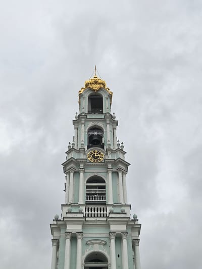 The image shows the Bell Tower of the Trinity Lavra of Saint Sergius in Sergiyev Posad, Russia. The tower is tall and ornate, with white and teal-colored facades, adorned with classical columns and decorative elements. A prominent golden clock face with Roman numerals is visible on one side of the tower. The top of the tower features a golden dome or spire. The sky is overcast and cloudy, suggesting it is daytime but with dull weather. The image is taken from a low angle, looking up at the structure. No people are visible in the frame, and the focus is solely on the architecture of the bell tower. The scene appears to be a tourist or religious site, given the historical and architectural significance of the Trinity Lavra.