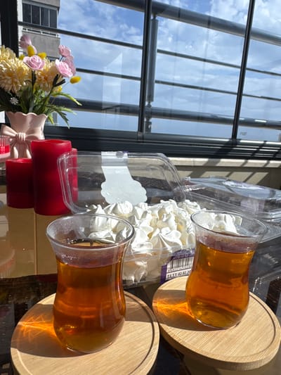 The image depicts two glasses of tea on wooden coasters, arranged on a reflective surface. Behind the glasses, a transparent plastic container filled with white meringues is visible. A red candle holder is positioned near the bottom of the frame, next to a small bouquet of artificial flowers. A large window with multiple panes dominates the background, offering a view of a cloudy sky and urban buildings, possibly indicating Erdemli, Turkey. The scene appears to be indoors, set against the backdrop of a city view through the window. There are no people in the image.