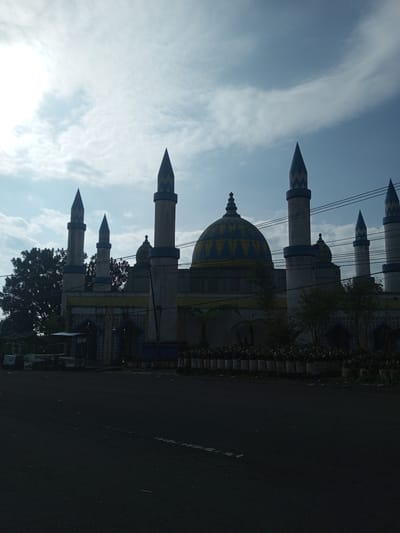 The image depicts a mosque in Mya Sit Kaing Ywar Thit, Myanmar. The mosque features a central dome with blue and yellow patterned decorations, flanked by several minarets with matching blue and yellow accents. A line of potted plants sits at the base of the mosque, near the road. The sky is overcast with patchy clouds. In the foreground is a paved road surface. A small structure with a canopy stands to the left of the mosque, and power lines are visible stretching across the sky. The overall scene appears dimly lit.
