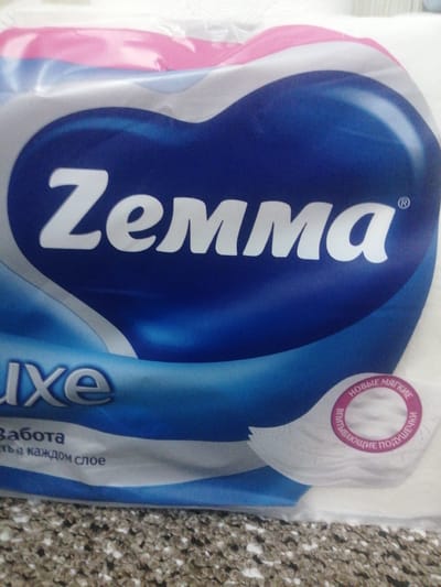The image shows a close-up of a package of "Zemma" brand toilet paper. The packaging is primarily blue and white with some pink accents. A large blue heart-shaped logo with the brand name "Zemma" in white lettering is prominently displayed. Below the logo, the word "luxe" is visible in silver lettering. Further down, in smaller text, the words "Забота" (meaning "care") and "ть в каждом слое" (meaning "in every layer") are visible. A circular emblem with pink text in Russian, reading "НОВЫЕ МЯГКИЕ ВПИТЫВАЮЩИЕ ПОДУШЕЧКИ" (meaning "new soft absorbent pads"), is located on the right side of the packaging. The packaging appears to be slightly crumpled. The background is out of focus but appears to be a countertop with a speckled pattern. There are no people or any indications of a specific location within the city of Achinsk, Russia, or any other urban environment. The image is an indoor product shot.