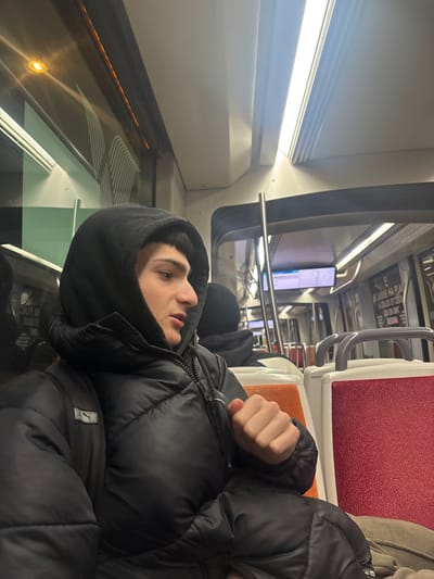 A person in a black puffy jacket and hood sits in a train car. The person's face is visible in profile, and their fist is clenched. Behind them, train seats are upholstered in red and orange. Above the seats, handrails are visible. The train car interior features a white wall with a window showing blurred scenery. Overhead lighting is present. Another passenger is sitting behind the main subject.
