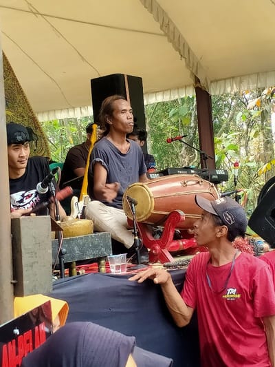 This image depicts an outdoor musical performance or rehearsal under a light-colored tent canopy in Tamansari, Indonesia.
In the center, a male musician, appearing middle-aged with long dark hair, sits cross-legged. He wears a dark blue t-shirt and light-colored trousers, and his hands are positioned over a large, reddish-brown kendang drum on a red wooden stand. A microphone is situated near the drum. Behind him to the right, a large black speaker cabinet is visible, and further back are two black electronic keyboards, one labeled "KORG" and another "Pa500," with an additional microphone on a stand.
To the left of the central drummer, a younger male musician, wearing a black t-shirt with a white graphic and a backward black baseball cap, sits. He holds a mallet over a rectangular wooden frame containing a small metal percussion instrument, possibly part of a gamelan ensemble. A microphone is also placed near this instrument. A patterned brown and gold fabric panel hangs vertically from a tent support pole to his left.
In the foreground on the right, another male individual, wearing a red t-shirt with a "TA HOMERGAL EST 2020" logo and a black baseball cap worn forward, leans forward. His right hand rests on a black-draped surface, and he looks up and to the left, observing the scene.
The performance area is draped in black fabric, with cables and equipment visible on the surface. A clear plastic cup containing liquid sits near the kendang drum. In the background, beyond the tent structure, lush green foliage and tree branches are visible, indicating an outdoor setting. Several other blurred figures are present in the background, including one behind the central drummer with a yellow electric guitar visible.