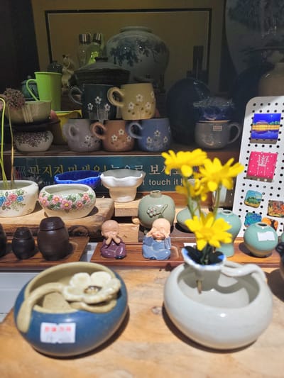 A shelf in a shop in Gyeongju-si, South Korea, displays a collection of ceramic items, primarily small pots and cups, alongside decorative figurines and a few yellow flowers in a vase. The collection is arranged on wooden shelves. In the foreground, a blue ceramic pot with a white flower ornament and a spoon sits on a wooden plank. To its right, a white ceramic vase holds a stem with a yellow flower. Behind these, a row of small, earth-toned jars and two monk figurines are visible. Further back, various ceramic cups with floral patterns in shades of green, blue, grey, and brown are stacked. There are also some larger ceramic vases and decorative plates on higher shelves. The overall impression is of a display of handcrafted ceramics, likely for sale.