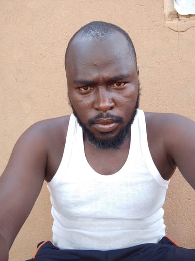 A man with dark skin, a shaved head, and a beard is wearing a white tank top. He is positioned in front of a textured beige wall. The man is facing the camera with a neutral expression. The lower portion of his torso and upper thighs are visible, showing dark-colored pants with an orange stripe. The scene is in the city of Mulke Barikin Sayejo, Nigeria.