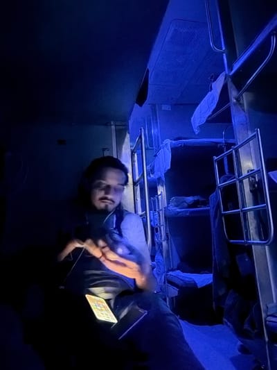 The image shows a man sitting in a dimly lit train compartment, illuminated by blue lights. He is wearing headphones and looking at his phone, which is resting on his lap. The compartment has bunk beds with blue blankets and white sheets. The man has a mustache and goatee and appears to be in his late twenties or early thirties. The scene suggests a journey on a train, possibly at night, given the darkness and the artificial lighting. The location is identified as Malya (Alot), India, a town in the Ratlam district of Madhya Pradesh, known for its railway station, which could explain the train compartment setting.