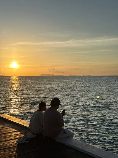 Two individuals, silhouetted against a vibrant sunset, sit on a wooden pier overlooking a tranquil ocean. The person on the left, a younger individual, is facing away from the camera, while the person on the right, an adult wearing a cap, is looking down at a mobile phone. Scattered on the wooden planks of the pier are a pair of white slip-on shoes. The setting sun casts a golden hue across the water, creating a shimmering reflection. In the distance, a faint outline of islands can be seen against the horizon. The sky transitions from a bright orange near the sun to a soft blue at the top. The overall scene suggests a peaceful evening at the beach in Baan Taling Ngam, Thailand.