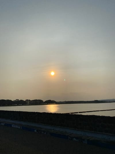 The image shows a serene natural landscape at dusk. The sun, a brilliant orb radiating light, is positioned in the upper left quadrant, casting a warm glow over the scene. The sky is a soft gradient of grays and yellows, indicating the transition from day to night. 

The foreground features a textured stone wall that runs along the right side of the frame. Beyond the wall, a calm body of water stretches horizontally, reflecting the soft light of the setting sun. On the far side of the water, a silhouetted treeline creates a dark, undulating horizon. The overall mood is peaceful and tranquil, evoking a sense of calm and quiet contemplation as the day ends. The location is identified as Bandungan, Indonesia, suggesting a tropical or subtropical environment. The image appears to be a still photograph, capturing a moment of natural beauty.
