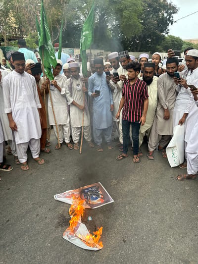 This image depicts a street protest in Karachi Division, Pakistan. A group of men, many wearing traditional Islamic attire, are gathered around a burning effigy on the asphalt road. Several individuals hold green flags with Arabic script. Some are observing the burning, while others are using their mobile phones. The background shows trees and buildings, typical of an urban environment. Smoke rises from the burning object, which appears to be a poster or flag. The overall scene suggests a demonstration with elements of anger or protest.