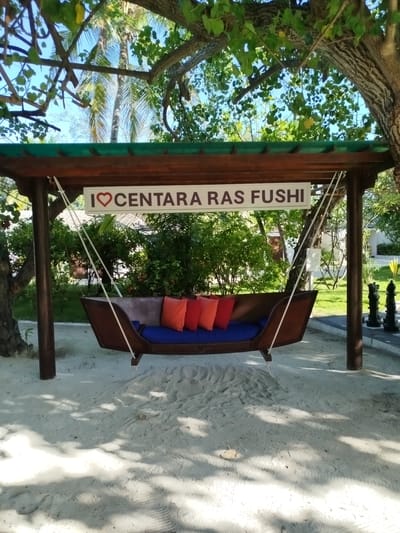 A wooden swing set with a boat-shaped seat hangs from a covered wooden structure on a sandy beach. The seat is adorned with four colorful cushions: two orange and two red. A white sign with red lettering that reads "I ❤️ CENTARA RAS FUSHI" is mounted on the underside of the structure above the swing. Palm trees and lush green vegetation surround the area, and the bright blue sky is visible through the leaves. In the background, there are hints of buildings and what appears to be a large outdoor chess set with black and white pieces. The scene is located on the island of Gulhi Falhu, Maldives.