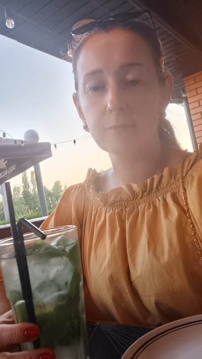 This is a selfie taken by a young woman outdoors, likely at a cafe or restaurant. She is wearing a light yellow, off-the-shoulder top with subtle gold chain detailing around the neckline. Her hair is dark and pulled back. She is holding a glass of iced green beverage, possibly a matcha latte or a mint drink, with a black straw. Her nails are painted a vibrant red.
The background suggests a casual outdoor setting, with what appears to be a brick wall and a dark wooden or metal overhang overhead. There are string lights visible, hinting at an evening or late afternoon setting. The sky is bright with a warm, hazy glow, suggesting either sunrise or sunset. Trees are visible in the distance, suggesting a natural element to the location. The overall mood seems relaxed and summery.
The word "BELGIAN LEGEND" is visible on a dark banner or umbrella in the background, providing a potential clue to the establishment's theme or origin. The image is oriented sideways, with the woman's face appearing near the top of the frame.