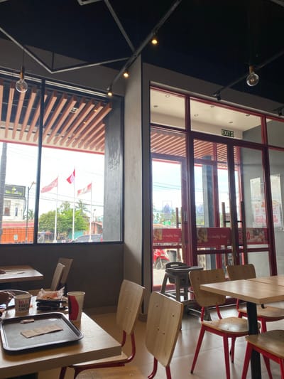 The image shows the interior and exterior of a KFC restaurant, likely located in Lhoksukon, Indonesia, as indicated by the text "Lhoksukon" visible on a street sign in the background. Inside the restaurant, several wooden chairs with red metal frames are arranged around tables. Some tables are empty, while others have remnants of a meal, including a red and white paper cup, a tray, and crumpled food wrappers. A sign inside the restaurant reads "KFC" and "It's Quality Time." There are also posters visible that mention "FREE" and "TO GO."
Through the large windows, the exterior scene shows a bright, sunny day with clear blue skies. Three flags on poles are flying outside, appearing to be red and white, possibly the Indonesian flag or similar designs. Trees and buildings line the street, suggesting a commercial area. The overall atmosphere appears casual and typical of a fast-food restaurant.
Visible text includes "EXIT" on a sign above a doorway, and brand names like "KFC" and "JAJAH." The "JAJAH" text, along with the overall streetscape, further points to an Indonesian location. The lighting suggests it is daytime. The image captures a blend of indoor dining and outdoor views, offering a glimpse into the typical environment of this KFC establishment.