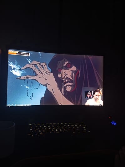 The image shows a computer screen displaying an anime scene alongside a small inset video feed of a person. The anime depicts a monstrous, dark-robed figure with glowing red eyes and sharp teeth, its skeletal hand reaching out. Below the anime, text in Cyrillic reads "х/ф D: Жажда крови," which translates to "Film D: Thirst for Blood." At the top left of the screen, "EME PARTY" is visible, along with a progress bar showing "519/1000." The inset video shows a man with brown hair, wearing a white tank top, looking towards the camera with a neutral expression. He appears to be in a dimly lit room, with a framed picture of a clown-like figure visible behind him. The computer keyboard below the screen is illuminated with yellow backlighting. The overall scene suggests someone is watching a movie or playing a game on their computer, possibly with a friend or during an online gaming session, in a dark room, likely at night given the ambient lighting conditions.