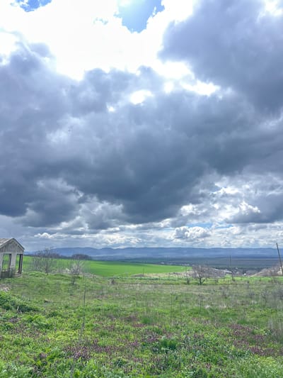 The image shows a landscape scene in Srednia, Bulgaria under a dramatic, cloudy sky. A field of green vegetation covers the right side of the frame, interspersed with patches of purple flowers. A wire fence runs through the field. In the foreground, a dilapidated concrete structure is visible. Beyond the field, there is a lighter green pasture and further back, a range of low mountains is discernible beneath the cloudy sky. The sky is dominated by grey storm clouds and patches of bright, sunlit clouds. Overhead power lines extend across the upper portion of the image.