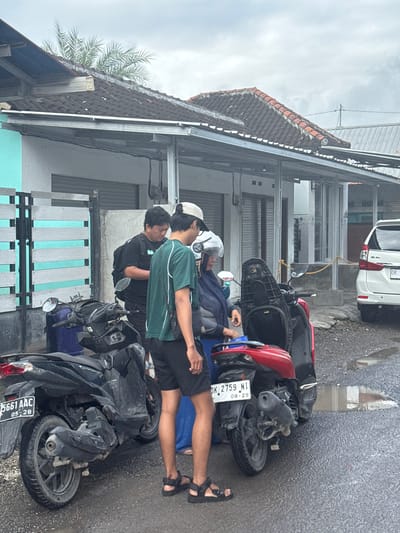 The scene shows three people in Kampung Toyapakeh, Indonesia, leaning on two parked motorbikes. One person is wearing a white helmet, a dark blue jacket, and sandals. Another person is wearing a green shirt, black shorts, and sandals. The third person is wearing a black shirt. The motorbikes are parked on a paved street. One is black, and the other is red. A white car is parked nearby. The street is lined with buildings. The sky is overcast.