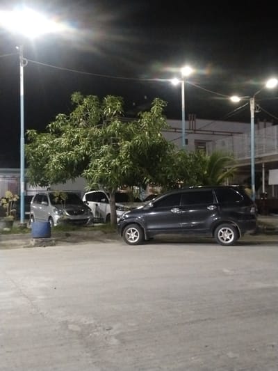 At night, a dark-colored SUV is parked on a paved surface in front of a large, leafy tree. Behind the tree, several other vehicles are visible, including a silver minivan and a white car. A building with an elevated walkway and railings is situated in the background. Streetlights illuminate the scene, casting a glow on the surroundings. The location appears to be a parking area in Keuniree, Indonesia.