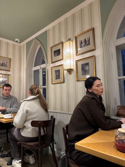 Three people are seated at a light-colored wooden table. One person wears a brown sweater and sits to the left, another person wears a light gray sweater, and the third person wears a gray sweater and sits in the bottom middle of the frame. The dining area has striped wallpaper, windows with arched tops, and framed pictures on the walls. A rectangular lighting fixture is mounted on the wall. A security camera is visible.
