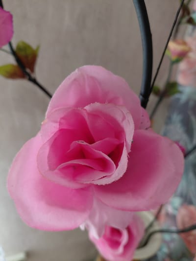 A close-up, eye-level shot focuses on a vibrant pink artificial rose. The rose is depicted with soft, layered petals that fade from a deep pink at the center to a lighter shade on the outer edges. A thin, black wire stem is visible to the right of the rose, curving upwards and out of frame. The background is a blurred, neutral tone, suggesting an indoor setting. The image does not contain any people, actions, or interactions, and there is no identifiable information to place it in the city of Новосёлово, Russia. The scene is static and emphasizes the texture and color of the artificial flower.