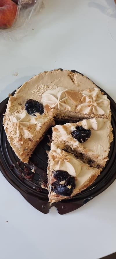 A light brown, round cake, likely with a coffee or caramel flavor, sits on a black plastic cake stand on a white surface. The cake has been sliced into several pieces, revealing layers of cake and frosting. It is decorated with swirls of white frosting, bits of what appear to be toffee or brittle, and several dark, whole cherries or berries. The overall impression is of a sweet dessert, possibly for a celebration or a treat. In the lower left corner of the image, a clear plastic bag contains several peaches, hinting at a possible fruit-themed gathering or meal. The lighting suggests it could be daytime, and the setting appears to be a home or indoor environment. No people are visible in the image. No text is visible.