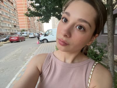 A young woman with brown hair and brown eyes is taking a selfie outdoors. She is wearing a pink tank top with a gold and white chain strap on her left shoulder. The background shows a street scene with several parked cars, two multi-story brick buildings, a white van, and a tree. One person appears to be working on the engine of a car in the middle ground. The scene suggests an urban residential area, possibly in Belozernyi, Russia, given the architectural style of the buildings and the types of vehicles.
