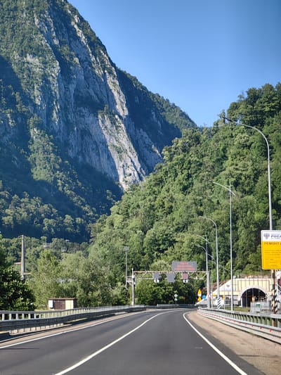 The image depicts a curved asphalt road with white lane markings, bordered by a silver metal guardrail on the right. The road leads towards a tunnel entrance, which is illuminated and appears to be made of concrete with arched openings. Several streetlights with curved poles are visible along the roadside. To the left of the road, a steep, rocky mountain face is covered with dense green trees. The sky is clear and blue. A yellow sign with Russian text and an emblem is posted on the right side of the road, indicating roadwork. Another sign with red text and an emblem is visible further down the road on the left, also indicating speed control. The scene suggests a mountainous region in Russia, potentially near Monastyr, given the context.