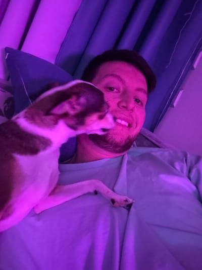 A man and a small dog are lying in bed. The man is on his back, smiling at the camera. The dog is standing on his chest, licking his face. The room is lit with purple and blue lighting. The man is wearing a light blue t-shirt. The dog is brown and white. The bed is covered with a blue sheet and pillows. The image was taken in Tokaryovo, Russia.