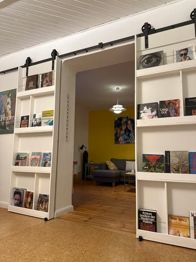 The image depicts an interior scene, likely a living room, with a doorway leading into another room. The primary focus is on two tall, white bookshelves flanking the doorway, which is fitted with a black sliding barn door track. The bookshelves are filled with various books, magazines, and artwork. On the left bookshelf, there's a large poster of a nude figure, and on the upper shelves, several books and magazines are visible, including one titled "mid-century modern complete." The lower shelves of the left bookshelf display more books and artwork.

The doorway opens into a room with yellow walls, a gray sectional sofa, a small side table with a candle, and a dark lamp. A modern pendant light hangs from the ceiling. The floor in both areas is made of wood planks.

On the right bookshelf, the upper shelves showcase framed artwork, including a close-up of an eye and portraits. Below these, books with titles like "Otto Dix" are displayed. The lower shelves on the right contain more books, including one with a painting of a ship and another titled "Knaurs Lexikon der Modernen Malerei."

The scene appears to be staged for a photograph, with no people present. The lighting suggests it is either daytime with indoor lighting or evening. The overall impression is one of a well-organized and art-focused living space. The mention of Ammeldingen bei Neuerburg, Germany, is not directly observable in the image itself and would require external information to confirm its location.